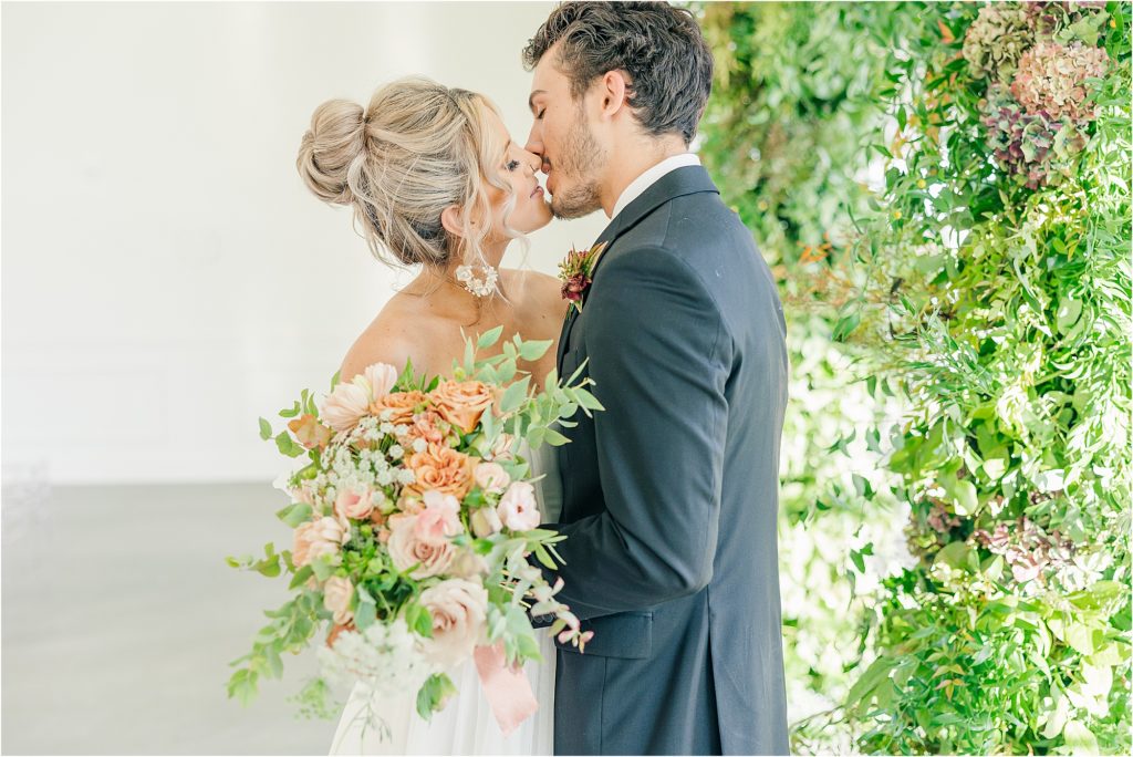 bride holding blush bridal bouquet kissing groom in black tux at Westwind Hills Westwind Hills ceremony with arched windows and greenery at alter Westwind Hills luxury wedding inspiration | Kelsey Alumbaugh Photography | #weddinginspiration #luxurywedding #stlouisweddingphotography #stlwedding #styledshootsacrossamerica