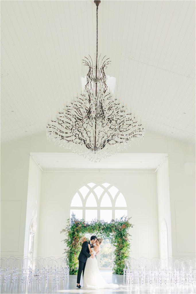 Bride and groom kissing in front of wedding alter Westwind Hills ceremony with arched windows and greenery at alter Westwind Hills luxury wedding inspiration | Kelsey Alumbaugh Photography | #weddinginspiration #luxurywedding #stlouisweddingphotography #stlwedding #styledshootsacrossamerica