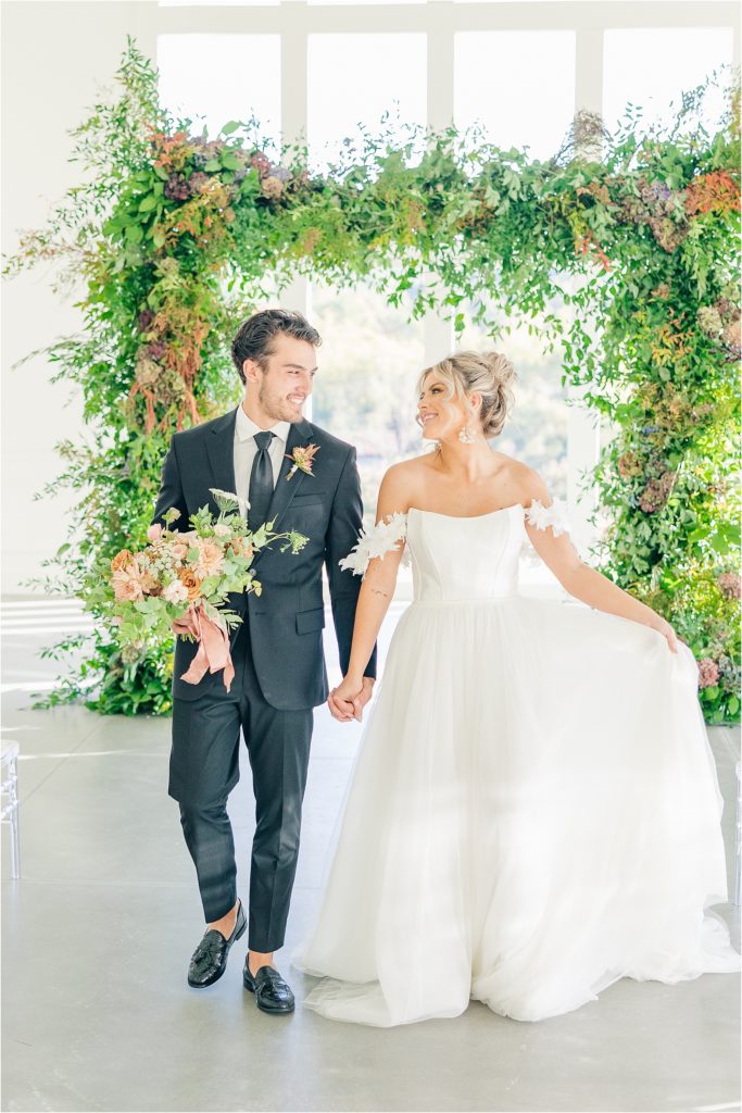 Groom in black tux holding blush bridal bouquet holding hands with bride in white off the shoulder wedding gown to walk down aisle Westwind Hills ceremony with arched windows and greenery at alter Westwind Hills luxury wedding inspiration | Kelsey Alumbaugh Photography | #weddinginspiration #luxurywedding #stlouisweddingphotography #stlwedding #styledshootsacrossamerica