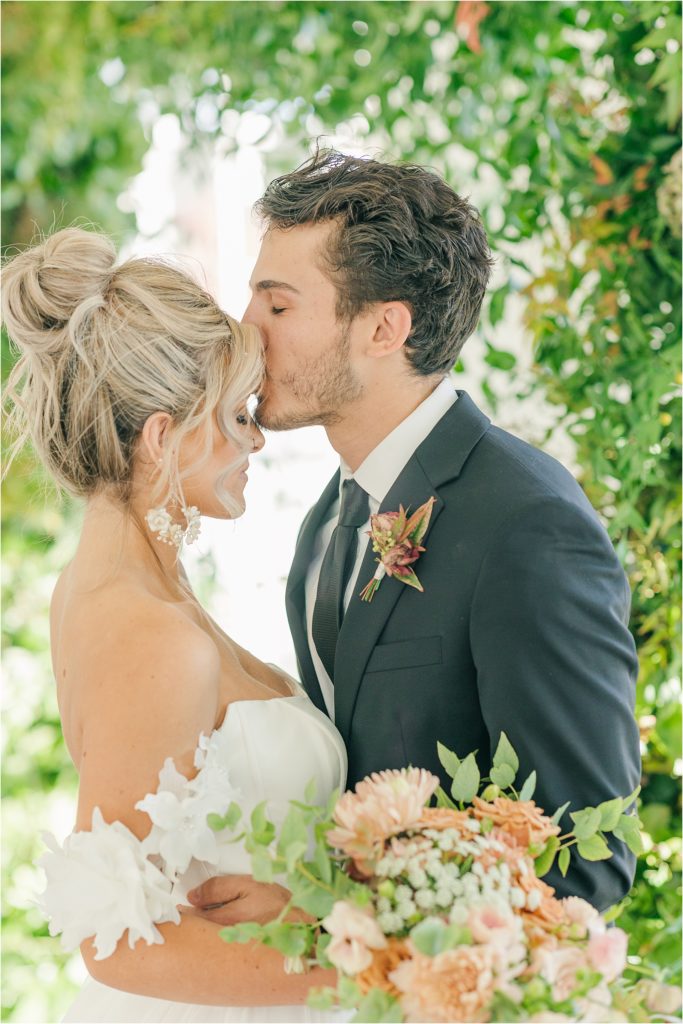 Groom in black tux kissing the forehead of bride in off the shoulder white wedding gown holding bouquet Westwind Hills luxury wedding inspiration | Kelsey Alumbaugh Photography | #weddinginspiration #luxurywedding #stlouisweddingphotography #stlwedding #styledshootsacrossamerica