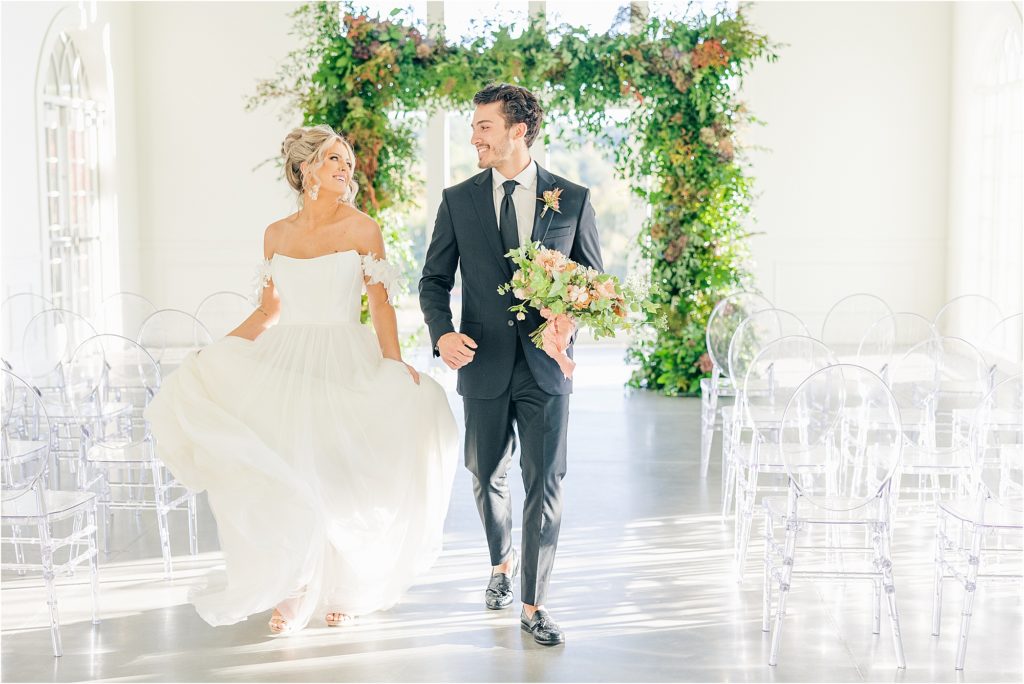 bride in white off the should wedding gown running down the aisle with groom in black tux holding blush and green bridal bouquet Westwind Hills luxury wedding inspiration | Kelsey Alumbaugh Photography | #weddinginspiration #luxurywedding #stlouisweddingphotography #stlwedding #styledshootsacrossamerica