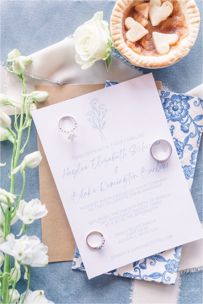 White wedding invitations on blue background with florals and rings and tiny apple pies Haylee + Blake | Missouri River spring wedding | Kansas City wedding photographer | Kelsey Alumbaugh Photogrpahy | #kansascitywedding #missouriweddingphotographer #missourispringwedding 